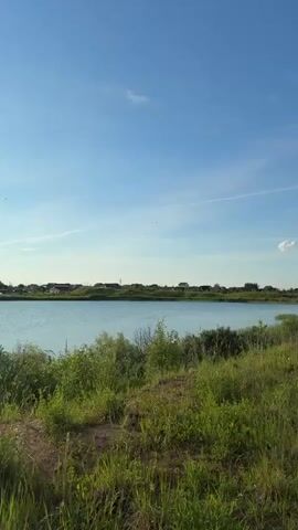 The video shows a rural landscape with a body of water on the left, and a dirt path on the right, leading through a grassy field. In the distance, houses and trees can be seen. The sky is blue with some clouds. There are a few people walking on the path in the distance. The camera is moving, giving a sense of walking through the scene.