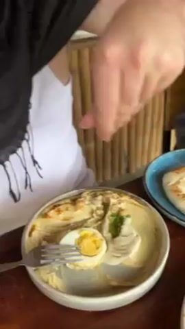 A person wearing a white t-shirt with a black scarf around their neck sits at a wooden table. They use their fingers to add a pinch of a red powder to a plate of hummus with a halved hard-boiled egg. A fork rests on the plate. A blue plate with what looks like pita bread is to the right of the hummus. A bamboo-like wall is in the background.