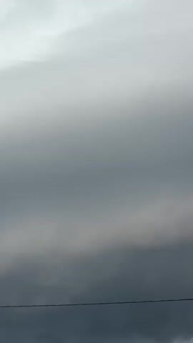 The video shows a view of the sky from below, looking upwards. The sky is filled with dark, ominous storm clouds. The clouds are heavy and appear to be moving. In the foreground, there are hints of buildings and trees, suggesting an urban or suburban environment. A power line is visible in the lower part of the frame, stretching horizontally. The lighting is dim, characteristic of a cloudy or stormy day. There are no people or overt actions visible, only the dynamic display of the weather.