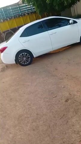 The video shows a white sedan parked on a dirt surface. The car is angled, with its right side facing the camera. The wheels are black with silver accents. The background includes trees, a yellow structure, and some buildings. The lighting suggests it is daytime. The video is short, lasting only five seconds.