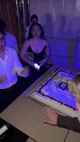 Here's a description of the video:

The video takes place in a nightclub setting. A rectangular cake decorated with the words "GRAM Gala Uptown" is the initial focal point.  People gather around the cake. The camera pans to show the inside of a crowded nightclub. Red balloons decorate the ceiling. The lighting is blue and red. People are standing, talking, and moving around the space.