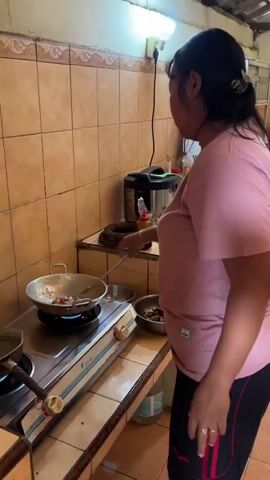 A woman is cooking in a kitchen. She is wearing a pink shirt and black shorts. She is standing in front of a stove, stirring food in a pan with a spatula. There are pots and pans on the stove, and a sink and cabinets in the background. The kitchen is tiled and has a fluorescent light overhead.