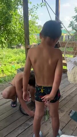 Two young boys are on a wooden porch. The boy in the foreground, facing away from the camera, is wearing patterned swim trunks. The boy in the background is crouched down, working with a black hose. Sunlight suggests it is daytime, and the lush greenery visible in the background indicates a warm season. The scene appears to be a casual, outdoor setting, likely a backyard or rural area, given the presence of trees, grass, and a hanging chair. There is no visible text in the media.