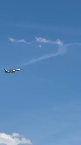 The media file captures an airplane in flight against a clear blue sky with a few wispy white clouds. The airplane is white with a blue and green tail fin, and it appears to be a commercial jet. A faint contrail is visible trailing behind the aircraft, curving slightly in the sky. The sun is bright, suggesting it is daytime, likely midday given the clear sky. The scene is set in an open sky, and while no specific geographical features are visible, the context provided places the location as Coslada, Spain. The media appears to be a still image or a short, unedited clip as there is no discernible action or event beyond the plane's flight. There are no people or text visible in the frame. The overall mood is one of clear skies and travel.