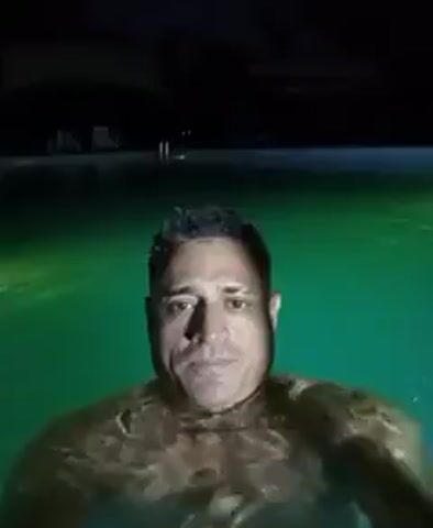 A man is in a pool at night. The pool water is illuminated, first by green light, then blue. The man is submerged to his chest, looking at the camera, and appears to be swimming.  He seems relaxed in the water. In the background, vague shapes suggestive of lounge chairs and structures are dimly visible in the dark.
