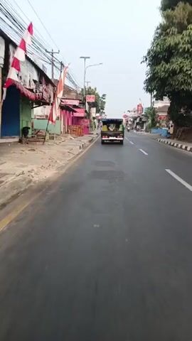 The video shows a view from a moving vehicle on a paved road. On the left side of the road, there are buildings with pink facades and awnings, some with signs that say "CRIA". There are also sidewalks and utility poles with many wires. On the right side of the road, there are more buildings and trees. A black pickup truck with a canopy is in front of the moving vehicle, carrying several people in green headscarves and possibly some cargo in the back. The truck is leaving tire marks on the wet road surface. A motorcycle is also visible in the distance on the right. The sky is overcast.