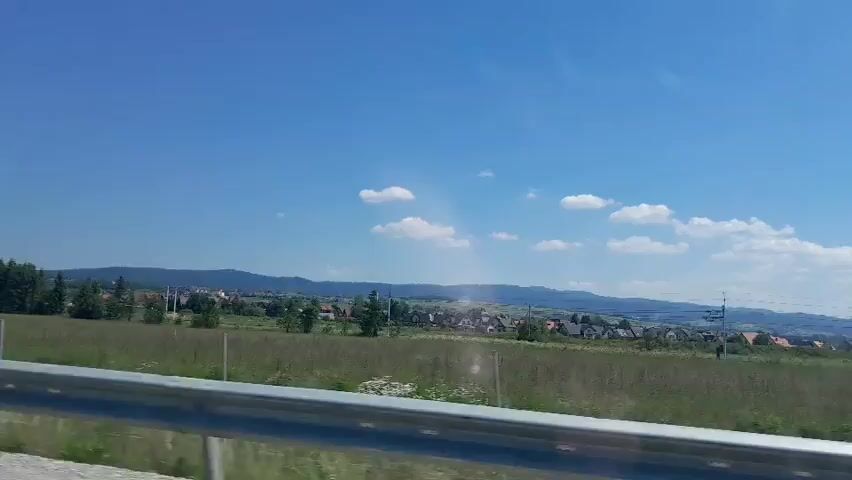 This video captures a scenic view from a moving vehicle, likely on a highway, looking towards a rural landscape under a clear blue sky with scattered white clouds. The time of day appears to be daytime, with bright sunlight illuminating the scene, suggesting fair weather.

In the foreground, a metal guardrail runs horizontally across the bottom of the frame, indicating the perspective of someone traveling by car. Beyond the guardrail, a wide, green field stretches towards a distant town or village. The field is covered in dry grass, suggesting a late summer or early autumn setting.

The middle ground is dominated by a cluster of trees, primarily coniferous, interspersed with residential buildings. These homes have red or brown roofs and appear to be scattered across the landscape, creating a picturesque village scene. Some individual houses are visible within the field as well.

In the background, a range of low, rolling hills or mountains forms a natural horizon. The overall impression is one of tranquility and natural beauty. The location is identified as Nowy Targ, Poland, which is consistent with a landscape featuring rolling hills and rural settlements. There are no people visible in the video, and no text is present. The activity is simply the movement of the viewer through this landscape.