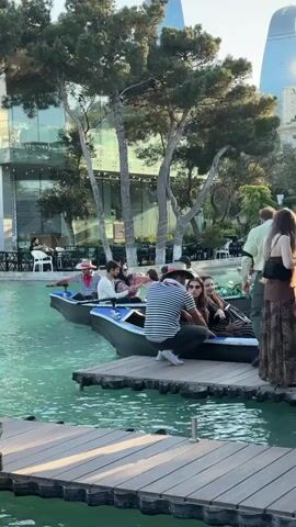 A wide shot captures a serene outdoor scene with a tranquil turquoise body of water. Two gondola-style boats glide on the water, each carrying passengers. The boat in the foreground, closer to the viewer, has two individuals seated inside. The boat in the background, slightly further away and to the left, carries three people, one of whom appears to be steering with an oar. The people in the boats are casually dressed, with some wearing hats. A wooden dock juts out into the water in the foreground. Behind the water, lush green trees with tall, slender trunks dominate the middle ground. Beyond the trees, a modern building with large glass windows and reflective surfaces stands tall, suggesting an urban or resort setting. The sky above is a soft blue, hinting at fair weather. The overall atmosphere is relaxed and picturesque.