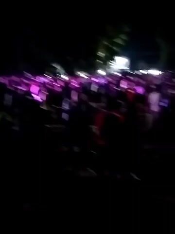 This is a video of a night event. There is a large crowd of people gathered in what appears to be an outdoor venue. In the background, there is a stage with bright lights and what looks like a band performing. There is also a large screen set up to the left of the stage, displaying an image of people. The overall atmosphere seems to be that of a concert or festival. The lighting is dim, with the stage and screen being the primary sources of illumination. The crowd is a dark silhouette against the brighter elements.