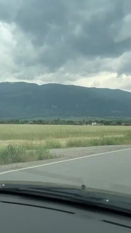 The video shows a view from inside a moving car, looking out towards a rural landscape. In the foreground, we see the dashboard of the car, partially obscured by the windshield. A gray asphalt road stretches out ahead, with white lines marking the lanes. To the left of the road, there's a field of tall, light green grass. Further into the background, a long, low building complex is visible, with a yellow facade and a red-tiled roof. Behind this building and to the left, a mountain range looms, its peaks shrouded in a green haze and partially obscured by a dense, overcast sky. The clouds are heavy and gray, suggesting an impending storm. As the car moves, a white van appears in the opposite lane, driving towards the camera, followed by a silver sedan.