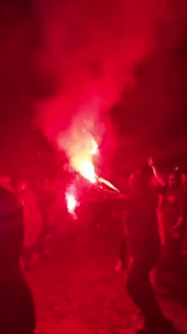 A group of people are gathered outdoors at night, illuminated by the bright red light and thick red smoke from a flare held by a person in the foreground. This individual holds the burning red flare aloft, waving it slightly. Other people are visible in the red glow and smoke, some dancing or raising their arms, with a few shirtless. The ground is dark, possibly dirt or grass. Towards the end of the video, a blue light briefly cuts through the red smoke, suggesting additional party lighting. The scene conveys a celebratory or party atmosphere.