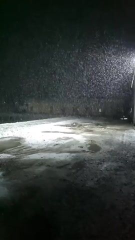 The video depicts a snowy scene at night. Heavy snowfall is visible under the light of a streetlamp. The ground is covered in patches of snow and puddles. In the distance, across a snowy bank, there is a body of water and a line of lights.