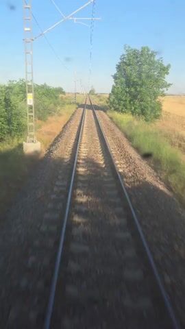 The video shows a view from a moving train on a sunny day. The camera is looking forward down a set of train tracks, which disappear into the distance. On either side of the tracks, there is vegetation, including trees and grass. Some of the fields appear to be harvested. There are power lines running overhead, supported by metal poles. The train tracks are ballasted with gravel, and the ties are visible. The motion of the train causes a slight blur in the foreground. The sky is clear and blue.
