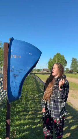 A young woman stands outdoors under a clear blue sky, next to a blue payphone booth mounted on a wooden post. She is wearing a plaid shirt and patterned pants. She holds the phone receiver to her ear and then hangs it back on the hook.