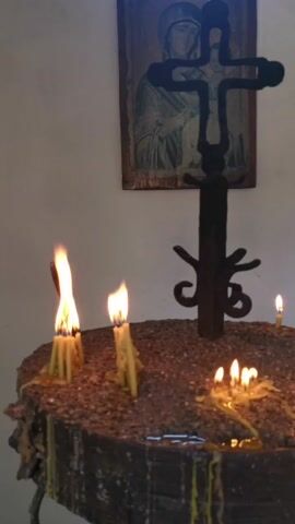 The video shows a close-up view of a candle stand with several lit candles. Behind the candle stand, a framed icon of the Virgin Mary is visible on a wall, and a black metal crucifix stands in the center of the candle stand. The candles are in various stages of burning, with some dripping wax. The flames are flickering and casting a warm glow on the surrounding objects. The environment appears to be dimly lit, suggesting it might be a place of worship or a prayer room. The video is static, with no camera movement.