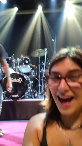 A man sings into a microphone on a concert stage under spotlights. He wears a dark cap, eyeglasses, a gray T-shirt, dark jeans, and sandals. Behind him, a drummer and another band member are playing instruments. The stage is pink. A hand wearing rings moves in front of the camera at times.