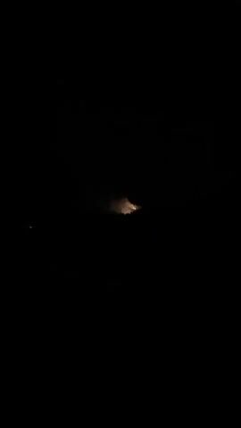 A dark video captures a distant fire at night. Flames and smoke are visible, illuminating the immediate surroundings with an orange glow. The fire appears to be on a hillside or elevated terrain, surrounded by darkness. There are no people visible, and the overall scene suggests a natural or industrial event occurring in a remote area.