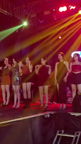 A group of six young women are standing on a stage in front of a large screen. The stage is lit with bright, colorful lights, including yellow and purple spotlights. The women are dressed in party attire, with various dresses and skirts. They appear to be posing or dancing, with some of them raising their arms or striking poses. The overall atmosphere is that of a performance or event.