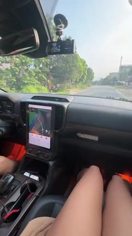 The video shows a first-person perspective from inside a car, likely in the front passenger seat. Two pairs of legs are visible, suggesting at least two occupants. The dashboard features a large, vertically oriented touchscreen display showing a navigation map with a route highlighted. A dashcam is mounted on the windshield, which also has two circular stickers with Vietnamese text, including "SÁO SÁO."

The car's interior is visible, with ambient orange/red lighting on the floor. A plastic Aquafina water bottle is seen on the passenger side floor. Outside the car, it's a bright, sunny day, with clear skies. The car is driving on a two-lane paved road. Trees line the sides of the road, and there are some buildings and power poles in the distance. Other vehicles, including cars and motorcycles, can be seen ahead on the road. The setting appears to be a rural or semi-urban area in Vị Xuyên Commune, Vietnam, given the architectural style and vegetation.