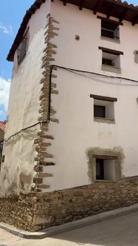 The video shows a close-up exterior view of a white stucco building with a stone foundation. The building has a tiled roof with exposed wooden beams. There are multiple small, square windows on the side of the building, some of which have wooden lintels. Some windows have planters with greenery. A few of the windows appear to be boarded up or have unfinished exteriors. Electrical wires are visible running along the exterior wall. The building is situated on a sloped paved street. In the background, a stone church tower with a clock is visible under a blue sky with scattered clouds. The architecture suggests a European setting, possibly a village or small town.
