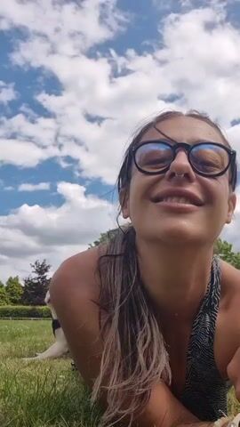 The video begins with a close-up shot of a woman wearing glasses and a nose ring, smiling and looking at the camera. She is outdoors, with a bright blue sky and white clouds visible in the background. Behind her, a light-colored dog with a purple collar is lying on the grass. The woman is lying down as well, with her head and shoulders in the foreground. The dog turns its head to the left and then back towards the woman. The camera angle shifts slightly, showing more of the dog and the grassy environment. The woman blinks and smiles again. The video is filmed in natural daylight.
