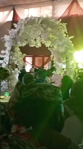 The video shows a gathering of people indoors, likely at an event venue with white draping and white floral decorations. People are seated, primarily facing a stage-like area, with some individuals standing and moving about. Most of the seated attendees are wearing head coverings. A person in a green and yellow patterned outfit is prominent as they stand in the aisle. The lighting is bright, but the overall image is somewhat grainy and obscured by the crowd. Upbeat music plays in the background.