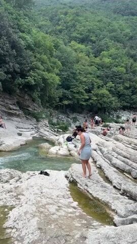The video captures a scenic view of a rocky riverbed with a group of people enjoying the water and surrounding landscape. A young woman in a light-colored dress is standing on a white, ridged rock formation near the edge of the water, with her back to the camera. She appears to be walking or posing. The river is a vibrant green and flows gently.

In the background, more people are scattered across the rocks and near the water. Some are sitting, others are standing, and a few are partially submerged in the river, suggesting they are swimming or wading. To the right, a man is seen bending over on the rocks.

The environment is lush and green, with dense trees covering a steep hillside that rises behind the river. The rocks are a pale gray or white, characterized by distinct horizontal ridging, indicating layers of sediment or erosion. The lighting suggests an overcast or diffused daylight, creating a soft atmosphere. The overall impression is one of a natural, popular spot for outdoor recreation.