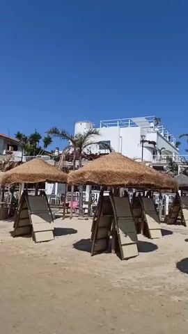The video shows a sunny beach with numerous thatched umbrellas providing shade over empty lounge chairs. A few people are visible in the background, mostly near the umbrellas. The beach is sandy, and a clear blue sky dominates the upper portion of the frame. Behind the beach, there's a white building with multiple levels and some lush green trees. The overall atmosphere appears relaxed and summery.