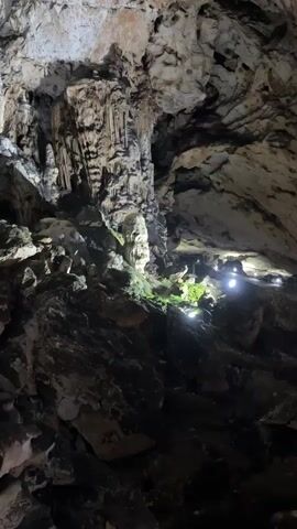 The video shows the interior of a cave with rocky formations and stalactites. The lighting is dim, but spotlights illuminate certain areas. The camera moves slowly, focusing on the texture of the rocks and the cave structure. An indistinct voice is heard.