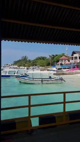 The video shows a pier with people walking around. The pier is covered with a corrugated metal roof. Underneath the roof are blue and yellow supports.  There is a railing along the side of the pier. The water is turquoise in color.  Several boats are visible in the water. In the distance, there is land with trees. The sky is blue with some clouds. There is a building on the pier. A person is standing near the edge of the pier.