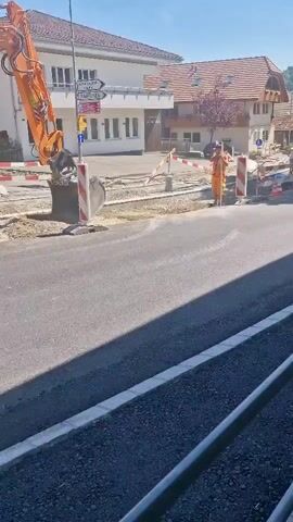 The video begins with a partial view of a red vintage convertible. The camera then pans to show the car driving away from the viewer down a paved road. The car, with its top down, has several passengers inside. The road appears to be under construction, with orange construction cones and barriers lining one side. An excavator is visible in the background. Buildings with traditional architecture and red tile roofs line the street, interspersed with trees. Further down the road, other vehicles and construction workers can be seen. The sky is clear and blue, indicating a sunny day. A person on a bicycle rides past the camera towards the construction area. The video ends as the vintage car continues down the road and out of frame.