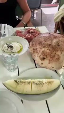 The scene is a table set with a variety of foods, including burrata with olive oil and black pepper, prosciutto, a large round loaf of bread, and a slice of melon sprinkled with what appears to be pepper. People are seated around the table, their arms and torsos visible. A partially-visible chair is in the background. The table has a white, slatted surface. The setting seems to be an outdoor dining area, judging by the daylight and glimpses of people walking by.