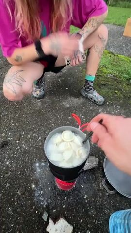 The video shows a close-up view of a pot with several pelmeni cooking in boiling water. A person's hand is visible holding a golden ladle, lifting some of the pelmeni out of the water to drain. In the background, another person is squatting down, wearing a pink hoodie and black shorts. They have tattoos on their legs and arms, and are wearing colorful socks and patterned clogs. The environment appears to be outdoors, with a grey, textured ground surface.