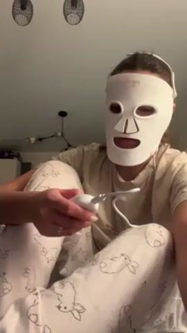 A woman wearing patterned pajamas and a white LED face mask sits cross-legged on what appears to be a bed or a couch. The mask is a rigid, white plastic or silicone with holes for the eyes, nose, and mouth. She holds a small white handheld device connected to the mask with a thin wire. She presses a button on the device, and the mask lights up with a red light, then presses the button again, and the mask lights up with a blue light. She then makes an adjusting motion to the mask on her face with both hands. The room is dimly lit, and in the background, there are two wall decorations resembling fans and a modern light fixture. A corner of what looks like a storage unit is also visible. The overall setting suggests an indoor residential environment.