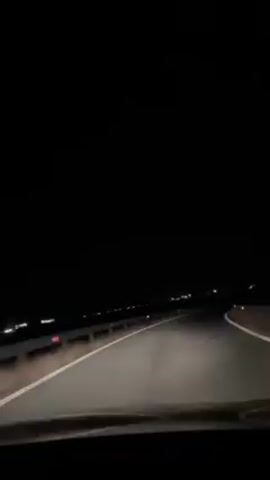A first-person perspective video shows a car driving on an empty, dark road at night. The road is asphalt with white lane markings and guardrails on the side. Distant lights are visible on the horizon. Car headlights illuminate the road ahead. Music is playing in the background.