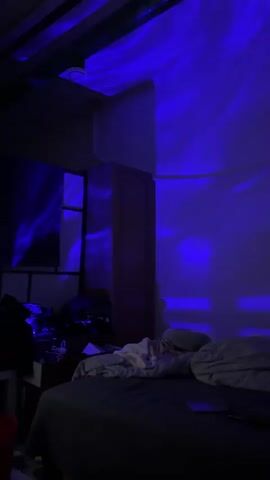 The video showcases an indoor room, likely a bedroom, bathed in dynamic, colorful projected light. The setting is dark, suggesting nighttime or a heavily dimmed room, creating an atmospheric ambiance.

Initially, from 0:00 to 0:01, the walls and ceiling are illuminated with flowing, wavy patterns of bright blue light. This quickly transitions from 0:01 to 0:03, where the projected light shifts to a vibrant mix of green and red, maintaining the same flowing, aurora-like patterns across the surfaces. Finally, from 0:03 to 0:05, the lighting changes once more to a blend of blue and purple/pink hues, continuing the swirling effect.

A bed with dark bedding and some rumpled lighter-colored sheets is visible in the foreground to the right. A tall, wooden wardrobe or cabinet stands against the wall on the right side of the frame, also catching the changing light. To the left, a surface reflects the projected colors, possibly a mirror or window, and some indistinct items are cluttered on a nearby surface. No people are present, and no specific activity is taking place other than the light show itself. The scene is set in Imatra, Finland.