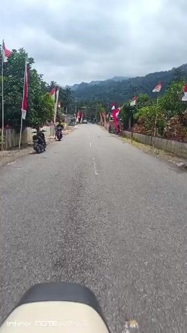 A perspective shot from a moving vehicle on a paved road with a white dashed line. The road is lined with trees, houses, and Indonesian flags. Several motorcycles and a car are visible on the road. In the distance, mountains are visible under a cloudy sky.