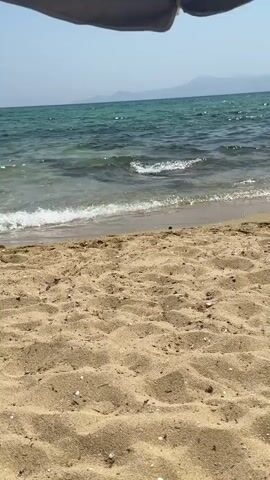A view of a sandy beach looking out at the ocean from under a partially visible umbrella. The water is clear with small waves lapping at the shore. The sand is a light brown and appears to be slightly wet near the water's edge. In the distance, a faint line of mountains can be seen against a clear sky. No people or specific objects are visible in the frame besides the umbrella.