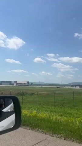 The video shows a view of an airfield with several large gray military aircraft parked on the tarmac in front of a large hangar. The view is from outside a chain-link fence, and there is a sign that says "Restricted Area Unathourized Entry Prohibited" attached to the fence. In the foreground, there is green vegetation. In the background, there is a mountain range on the horizon and a blue sky with scattered white clouds. The camera appears to be positioned inside a vehicle. The car's side mirror is visible on the left.
