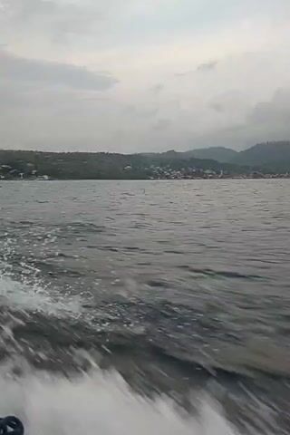 A boat is moving through the water creating a wake. In the background, there is land with hills and buildings. The sky is cloudy.