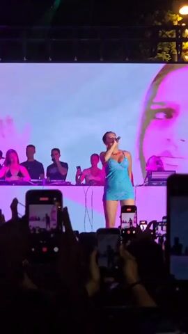 The video shows a female singer on stage performing in front of a large screen displaying abstract visuals. She is wearing a blue dress and holding a microphone. Behind her, several people, some appearing to be DJs, are visible on a raised platform. The stage is lit with colorful lights, including green and purple spotlights. In the foreground, the hands of audience members holding up mobile phones to record the performance are prominent. The overall environment appears to be an outdoor concert or festival at night, with trees and lighting visible in the background.