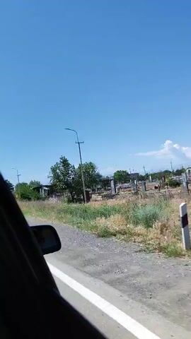 A camera mounted in a vehicle records a view from the passenger side window as the vehicle travels along a road in daylight. The road is asphalt with a solid white line on the right side. The vehicle is moving, and the landscape is visible outside the window. A rearview mirror is partially visible in the bottom left corner of the frame, showing a reflection of the vehicle's interior. Beyond the road, there's a strip of dry, grassy ground and scattered trees. In the background, there are some buildings or structures, appearing somewhat dilapidated or under construction, and more trees. The sky is bright blue with a few distant clouds. The overall scene suggests a rural or semi-rural environment on a clear, sunny day.