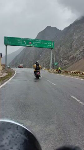 The video shows a motorcycle rider from a first-person perspective driving on a wet, paved road in a mountainous region. The rider is moving forward, away from the camera. In front of the rider, there is another motorcycle with a rider wearing a yellow backpack and a helmet. A green overhead sign indicates distances to various locations including Leh (325 km), Keylong (78 km), and Atal Tunnel Rohtang (57 km) and Manali (100 km). The sign also mentions the Border Roads Organisation and Project DEEPAK. Cars and a truck are visible on the road in the distance. The sky is overcast, and the mountains are covered in mist. The road surface is glistening, indicating recent rain.