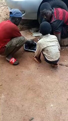 Three individuals are visible in the video, likely two adults and one child, working on a car outdoors. The car, silver in color, is positioned with its underside exposed. The individual on the left is wearing a red shirt, beige shorts, a blue baseball cap, and red sandals. He is squatting and appears to be examining or working on a black rectangular object, which is resting on a piece of cardboard on the ground. Another man, wearing a red and black striped shirt and dark pants, is squatting behind and to the right of the child, looking down at the object. The child, wearing a checkered shirt and dark pants, is also squatting, looking at the same object. The ground is dirt. There is a tire in the background. The three individuals appear to be collaborating on a mechanical task related to the car.