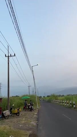 A vertical video captures an overcast sky above a rural road lined with tall grass and utility poles. On the left, a person is seated next to a motorcycle with a yellow storage box. Another person is standing, and two more are seated, beneath a green umbrella. Several motorcycles are parked on the grassy shoulder. The road curves gently to the right and recedes into the distance, with a few motorcycles visible on it. A mountain range is faintly visible in the background.