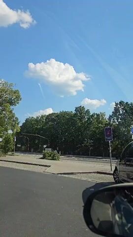 The video shows a view from inside a vehicle, looking forward and slightly to the right. The sky is blue with several white clouds, one of which is large and fluffy. A line of trees forms a backdrop across the scene. In the foreground, the edge of the vehicle's rearview mirror is visible on the right side. A road with grey pavement and a white curb is in front. To the left, a traffic signal with red lights illuminated is visible. Further to the right, a sign with a purple background and the word "TAXI" in white text is mounted on a pole. A dark-colored car is parked to the right of the taxi sign. The overall scene suggests an outdoor urban or suburban environment on a clear day.