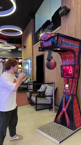 A man is playing a Spider-Box arcade game. The game is red and black and decorated with images of Spider-Man. The man is wearing a white t-shirt and gray shoes and is punching the punching bag on the game. The score on the game is 56.9. The game is located in an arcade or entertainment center, with couches and other arcade games visible in the background.  A woman walks behind the punching game.