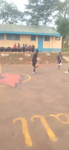 The video shows two brief scenes of people playing basketball on outdoor courts. In the first scene, two men face off, with one dribbling the ball while the other defends. The second scene shows multiple players on a different court. One player in a yellow jersey with the number eight drives to the basket and shoots over a defender in a navy blue jersey.  The shot goes in, and the player in yellow raises his arms in victory. The court is brown and reads "St. Mary's West" in yellow letters.  A simple basketball hoop and backboard are present. There are trees, buildings and other people in the background. The sky is overcast.