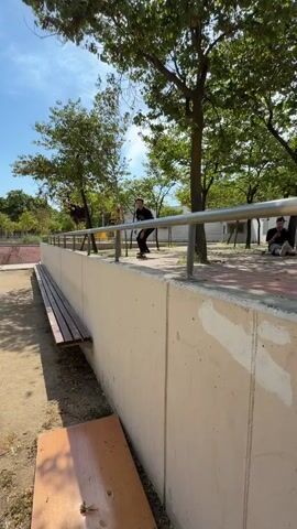 A young person with short dark hair, wearing a black t-shirt and black pants, is skateboarding in an outdoor park. The person is performing a trick on a metal railing, balancing on their skateboard. In the background, there are trees, a concrete wall, a park bench, and buildings. Another person is sitting on a bench in the background. The skateboarder successfully completes the trick and lands on the ground. The video captures the skateboarder approaching the railing, grinding on it, and then jumping off. The scene is well-lit by sunlight.