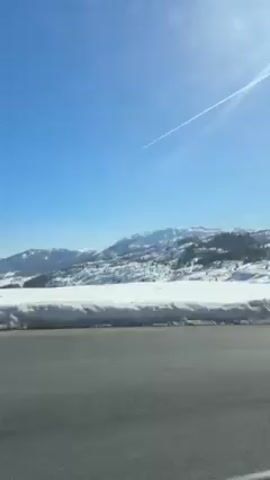 The video shows a road next to a snow-covered landscape under a clear, blue sky. A jet contrail crosses the sky. A road sign is visible on the right side of the road. The landscape includes snow-covered mountains. The sun shines brightly. There is someone speaking in the background, and music is also playing.